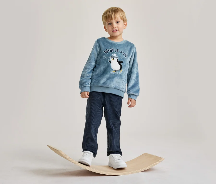 Jeune garçon debout sur une planche d'équilibre en bois. Il porte un haut bleu à manches longues avec un motif de pingouin et le texte « WINTER FUN », un pantalon en velours côtelé bleu foncé et des baskets blanches.