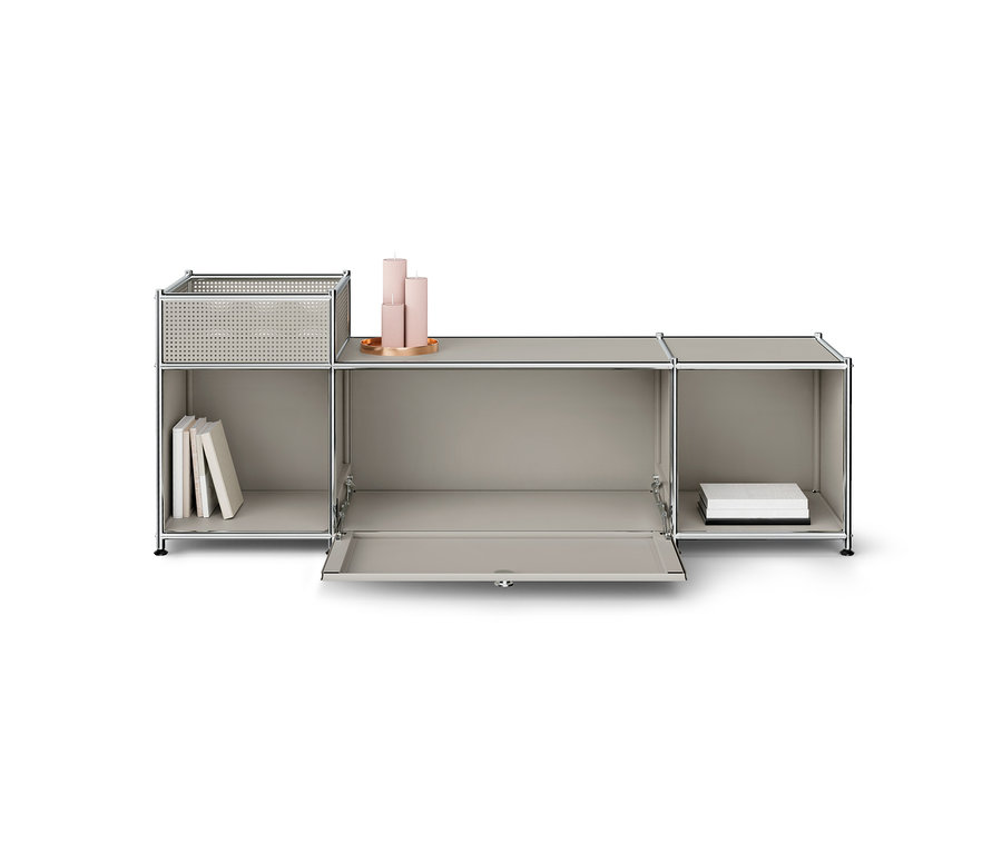 Meuble de rangement modulaire gris clair avec cadre chromé. La section gauche a une étagère ouverte avec des livres et un panier perforé sur le dessus. La porte abattante de la section centrale est ouverte, et trois bougies roses sont posées sur sa surface supérieure. La section droite est une étagère ouverte avec des boîtes blanches.