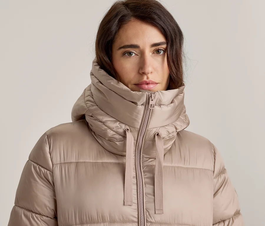Gros plan d'une femme aux cheveux foncés portant une doudoune marron clair. La veste a un col haut et rembourré avec une fermeture éclair et des cordons visibles, et elle regarde directement l'appareil photo.