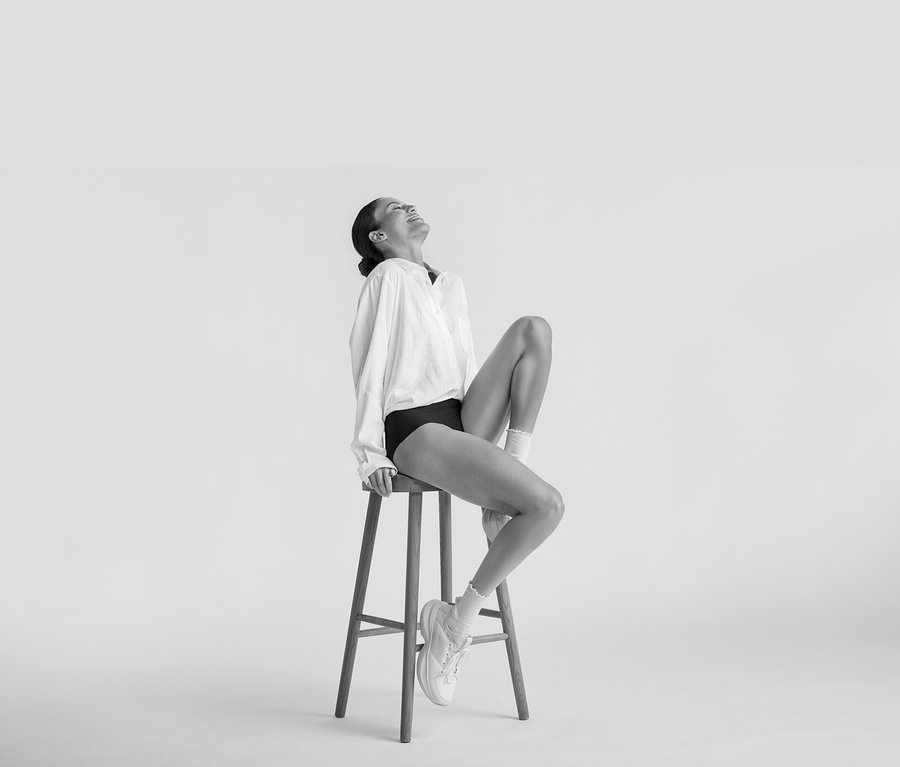 Femme assise et souriante sur un tabouret, portant une Blouse longue en lin blanche, des sous-vêtements noirs, des chaussettes blanches et des baskets blanches.