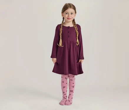 Une jeune fille aux tresses blondes sourit en se tenant debout, vêtue d'une robe prune à manches longues avec des boutons et des poches, et d'un collant rose à motif floral foncé.