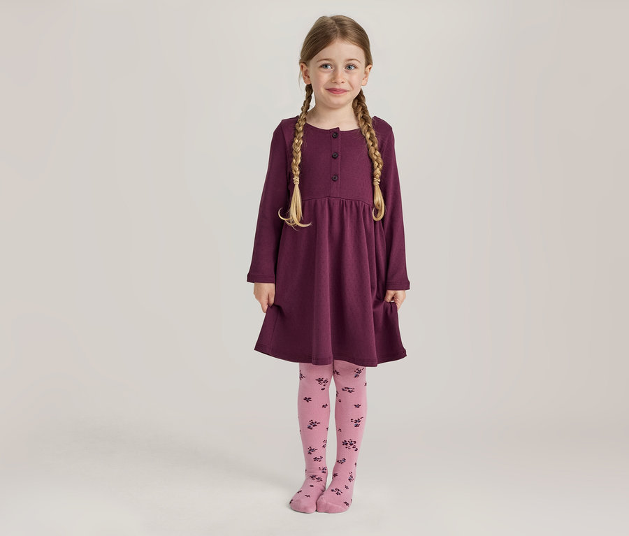 Une jeune fille aux tresses blondes sourit en se tenant debout, vêtue d'une robe prune à manches longues avec des boutons et des poches, et d'un collant rose à motif floral foncé.