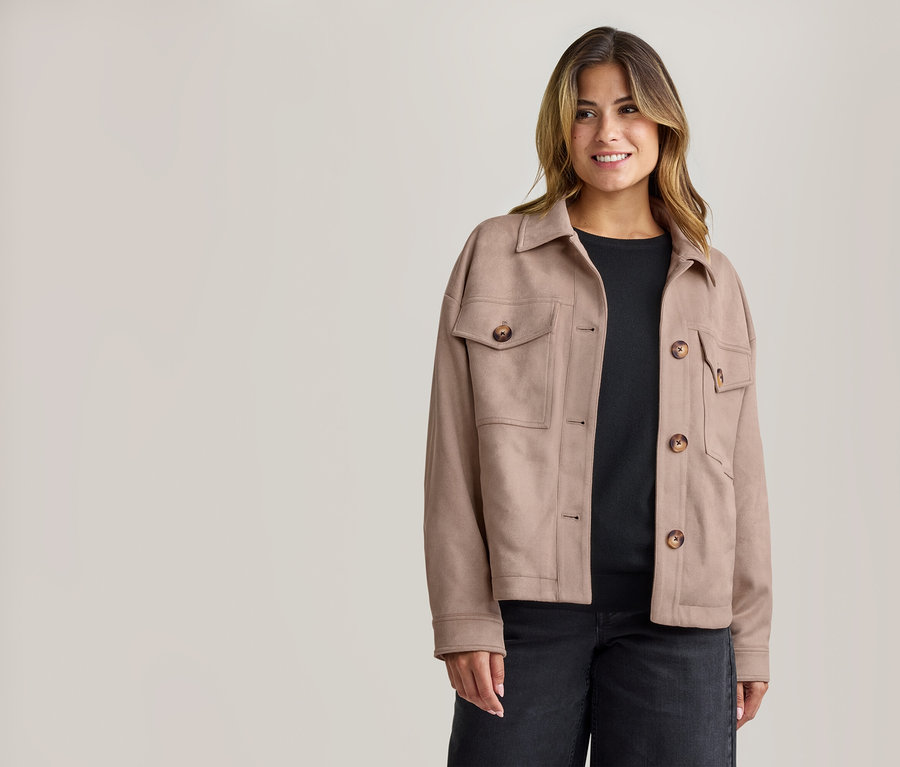 Femme souriante portant une veste beige à col avec des boutons marron et des poches, un haut noir et un jean foncé.