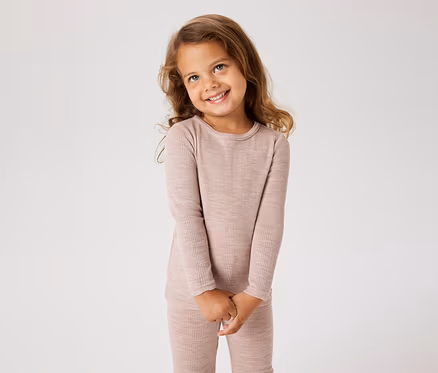 Jeune fille aux longs cheveux bruns, souriante et regardant vers le haut, portant un haut côtelé à manches longues rose clair et un pantalon assorti, les mains jointes.