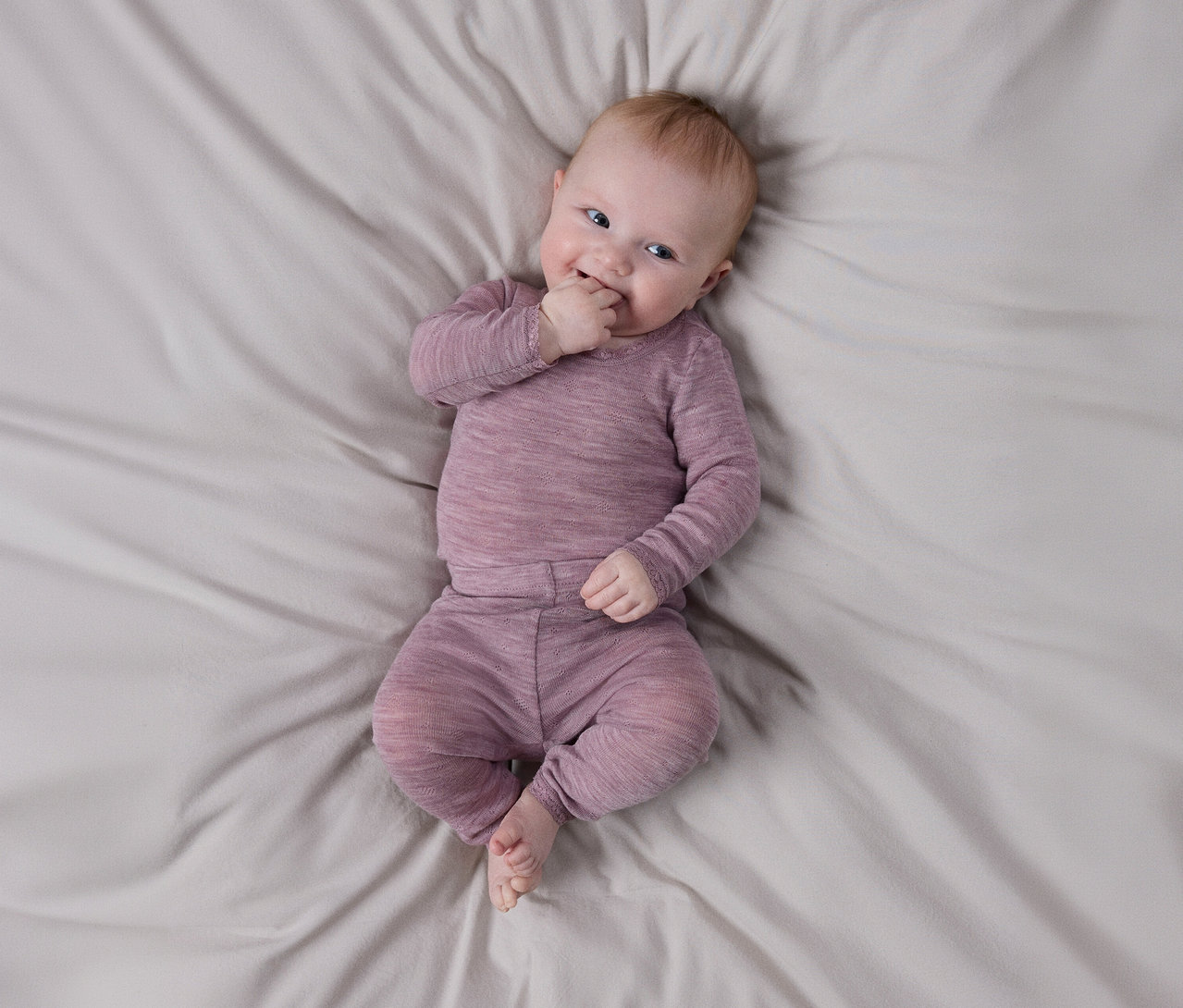 Bébé souriant allongé sur un drap gris clair, portant un haut violet texturé à manches longues et un pantalon assorti, une main près de sa bouche.
