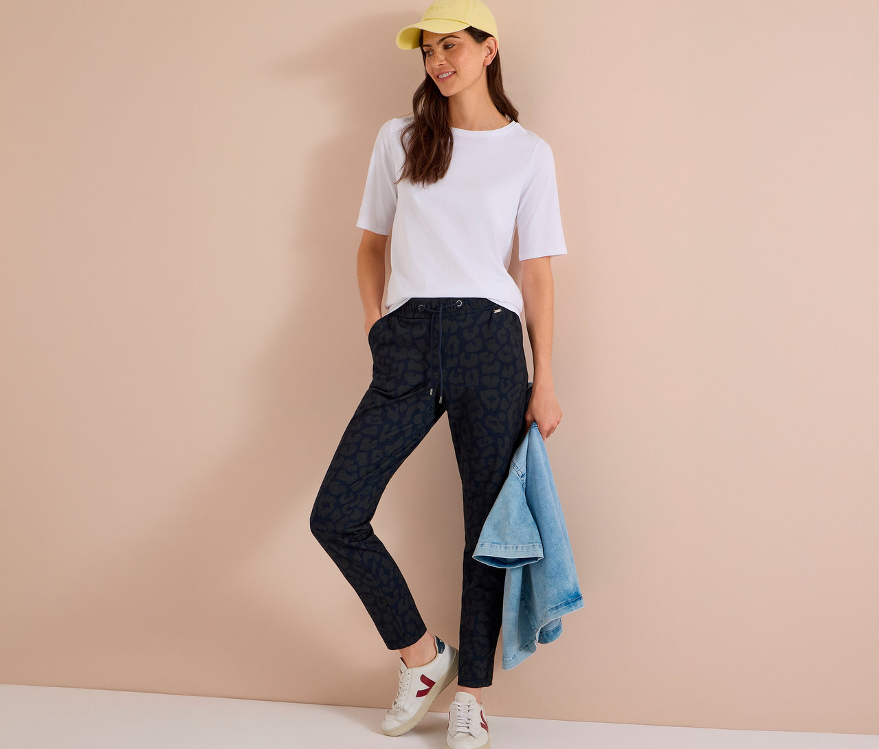 Femme portant un t-shirt blanc, une casquette jaune, des baskets blanches et un pantalon en jersey CECIL imprimé, tenant une veste en jean.