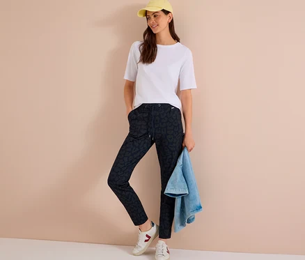 Femme portant un t-shirt blanc, une casquette jaune, des baskets blanches et un pantalon en jersey CECIL imprimé, tenant une veste en jean.