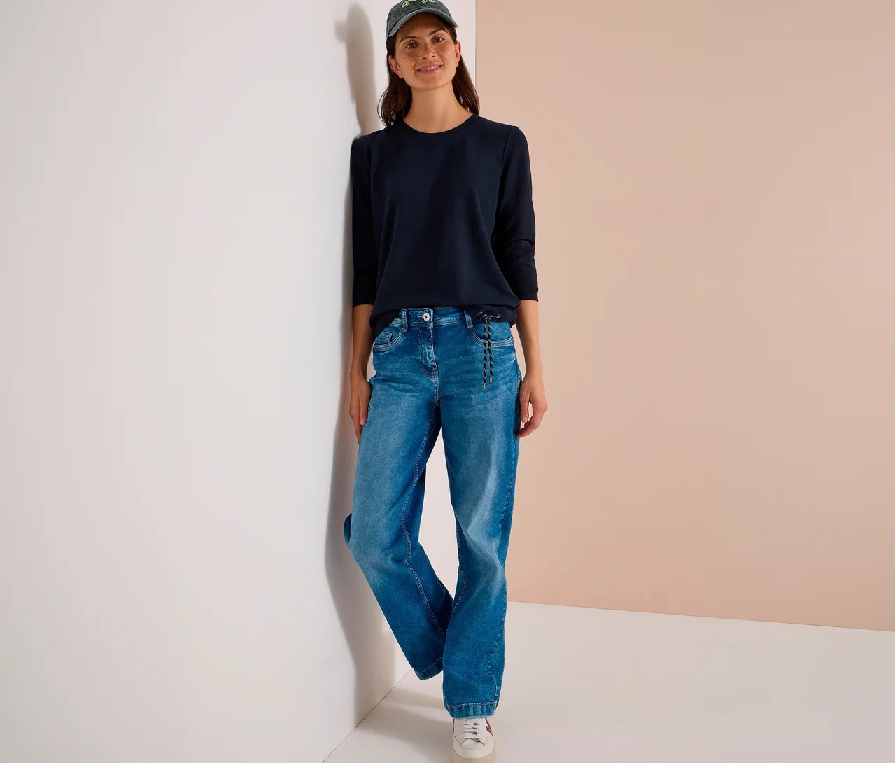 Une femme souriante s'appuie contre un mur, vêtue d'un T-shirt CECIL bleu foncé, d'un jean bleu, de baskets blanches et d'une casquette de baseball verte.