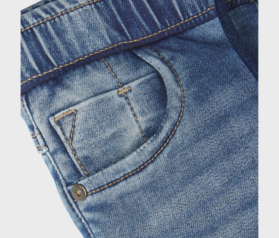 Gros plan d'un jean slim sweat bleu NAME IT, avec taille élastique, poche avant, petite poche et bouton métallique estampillé « NAME IT ».