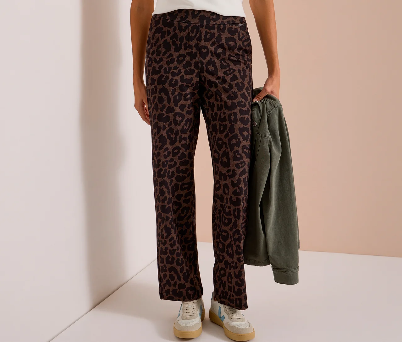 Personne portant un pantalon en tissu CECIL à imprimé léopard sur toute la surface et tenant une veste verte.