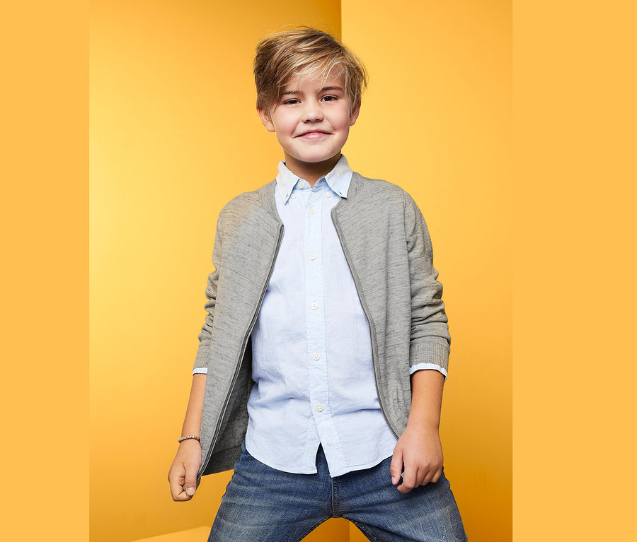 Garçon posant sur fond jaune en veste en maille, chemise bleue et jean.