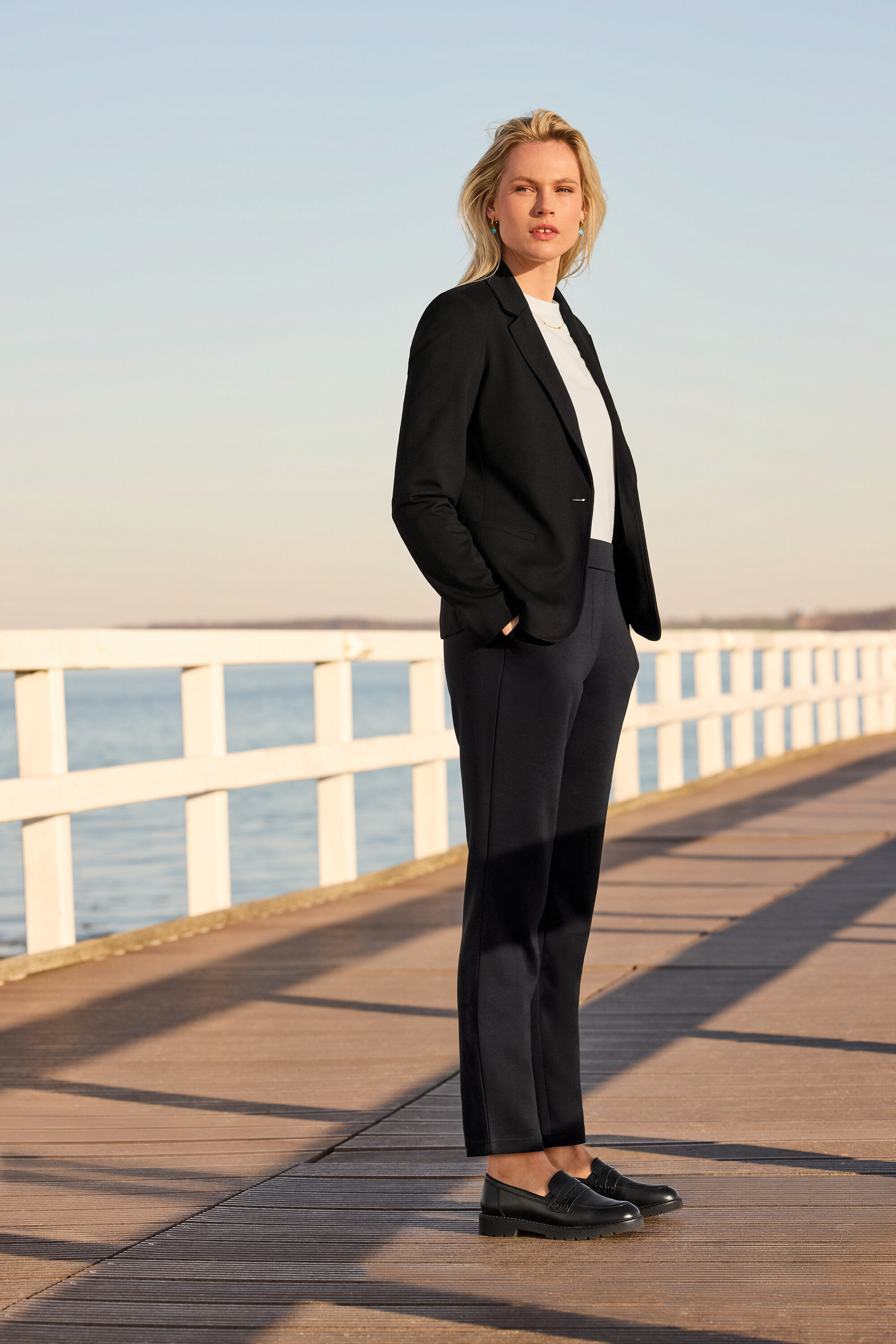 Une femme debout sur une jetée porte un pantalon de jogging, un T-shirt basique blanc, un blazer en jersey et des mocassins en cuir. Elle porte des créoles avec pendentif et une chaîne à navettes.