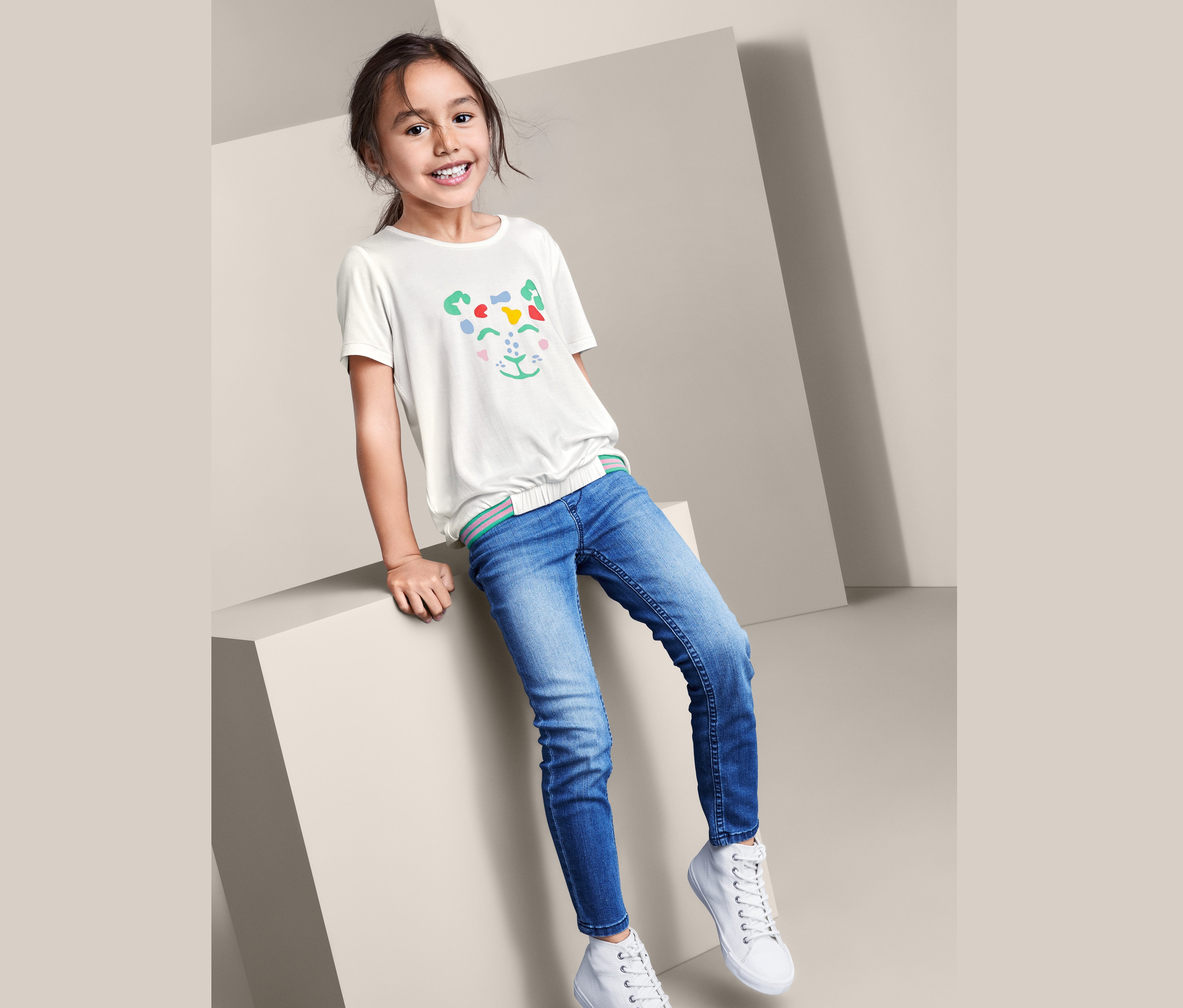 Une fille est assise sur un cube et porte un jean, des baskets blanches et un t-shirt à enfiler blanc avec un visage d'animal.