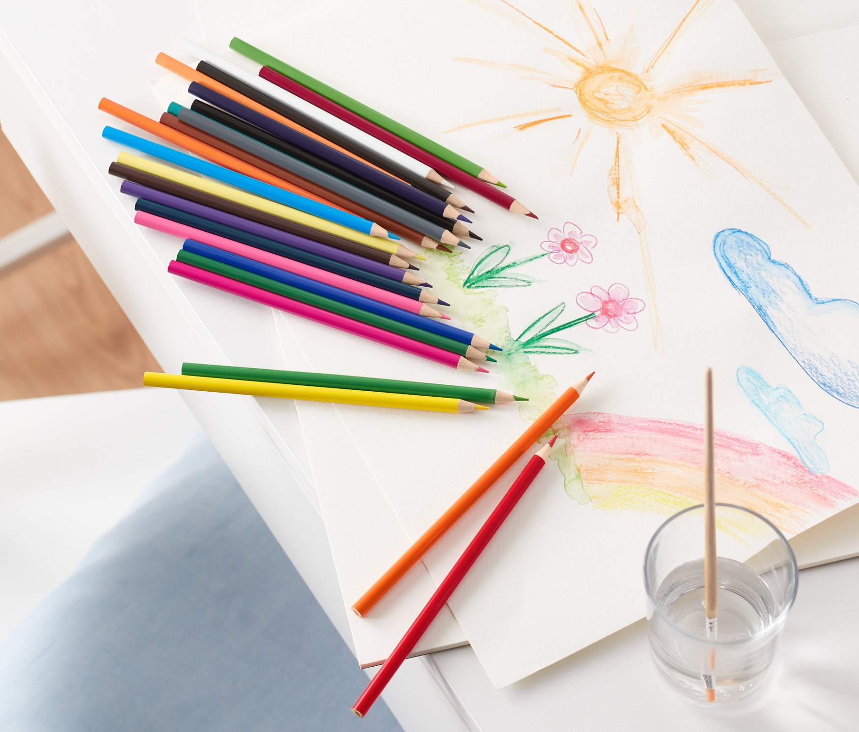 Sur une table blanche reposent un dessin de soleil, de fleurs, de nuages et d'un arc-en-ciel, à côté de crayons de couleur aquarelle et d'un verre avec de l'eau et un pinceau.