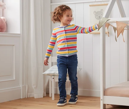 Une fille se tient dans une pièce et touche des décorations sur un cadre de lit. Elle porte une jaquette en maille, à rayures dans les couleurs d'automne.