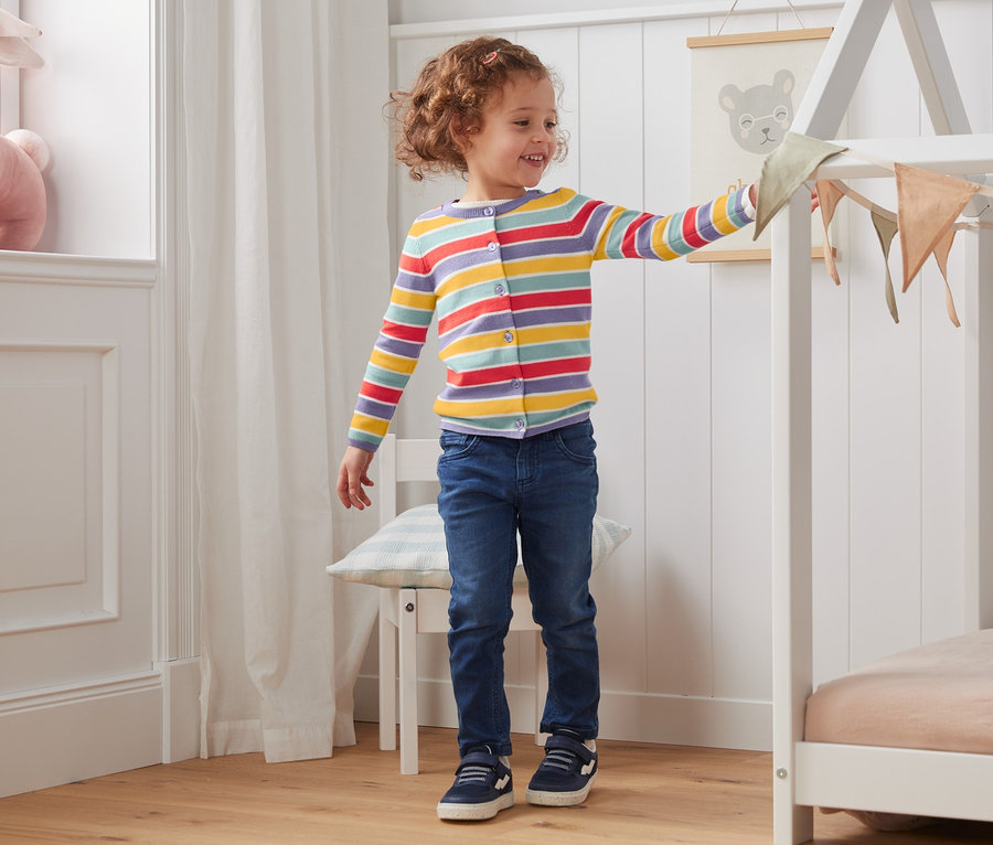 Une fille se tient dans une pièce et touche des décorations sur un cadre de lit. Elle porte une jaquette en maille, à rayures dans les couleurs d'automne.