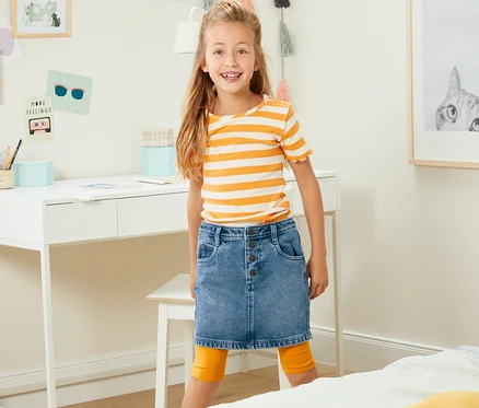 Fillette souriante posant en jupe en jean devant un bureau.