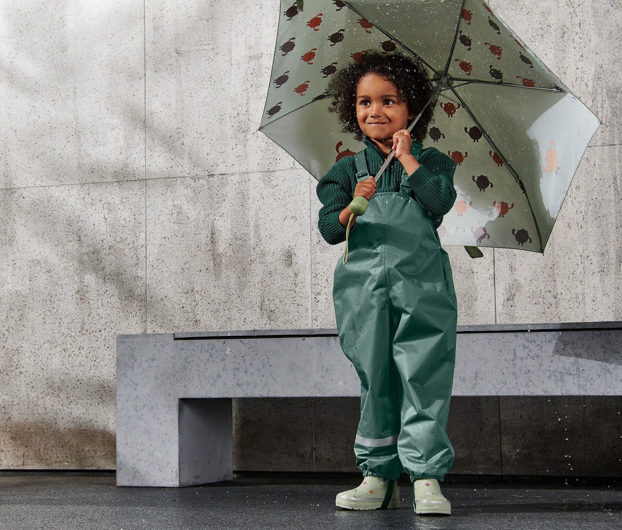 Fille tenant un parapluie à motif de tortues. Elle porte un pantalon de pluie thermique pour enfant, vert foncé.