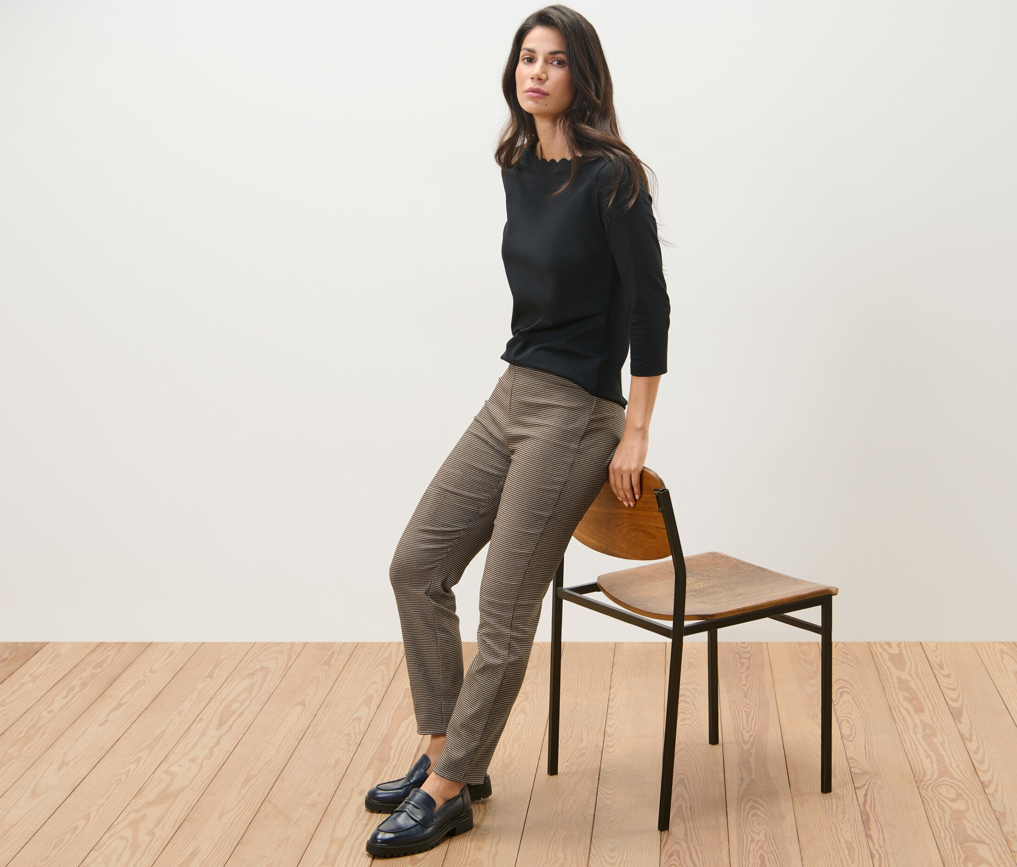 Une femme est assise de côté sur une chaise, portant un haut noir et un pantalon stretch avec jacquard pied-de-poule.