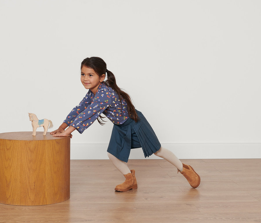Fillette s'appuie sur une table avec un cheval en bois, portant une chemise fleurie bleue et une jupe kilt bleu marine pour tout-petits.