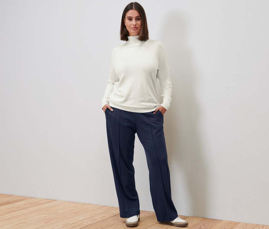 Une femme pose, portant un pull à col roulé en maille fine blanc crème, un pantalon en jersey bleu foncé avec nervure et des baskets en cuir.