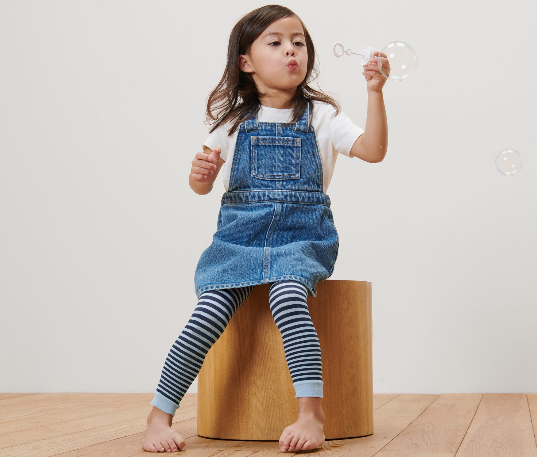Une fille est assise sur un tabouret en bois et fait des bulles. Elle porte une robe en jean et 2 leggings fins pour enfants.