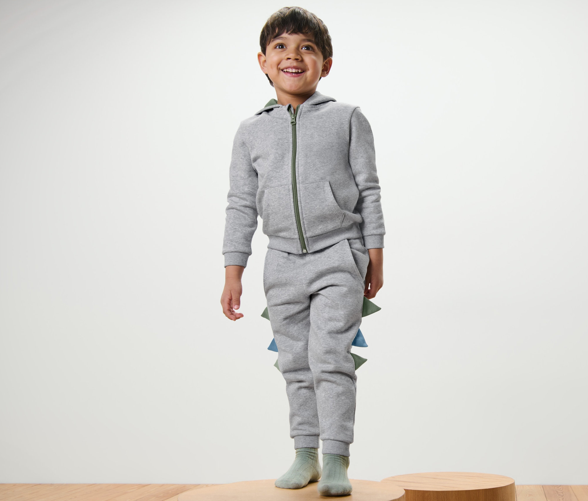 Un garçon se tient sur un socle en bois et porte un pantalon en sweat pour enfant avec application dinosaure gris.