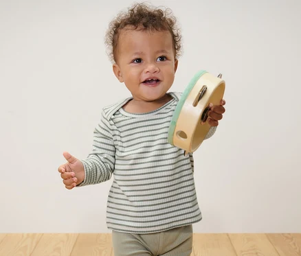 Un bébé debout tenant un tambourin. Il porte 2 t-shirts pour bébé à manches longues avec structure côtelée, bleu clair et vert.