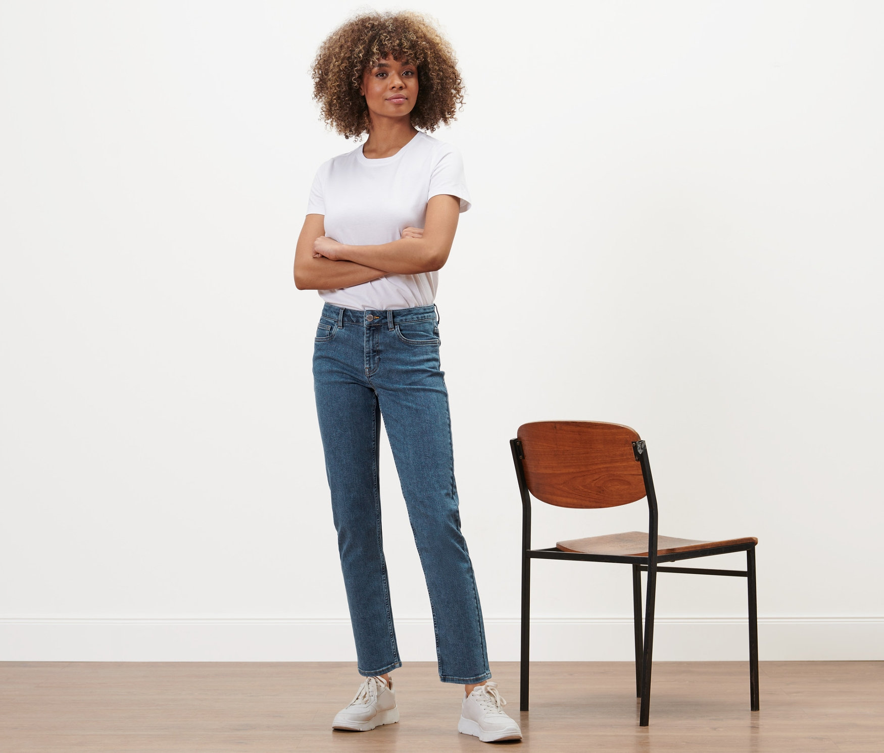 Une femme aux bras croisés se tient à côté d'une chaise et porte un jean droit en denim « Fit Juna », bleu moyen.