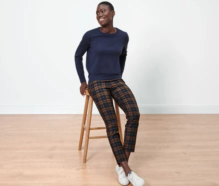 Une femme noire est assise sur un tabouret, portant un pull bleu et un pantalon stretch à carreaux.
