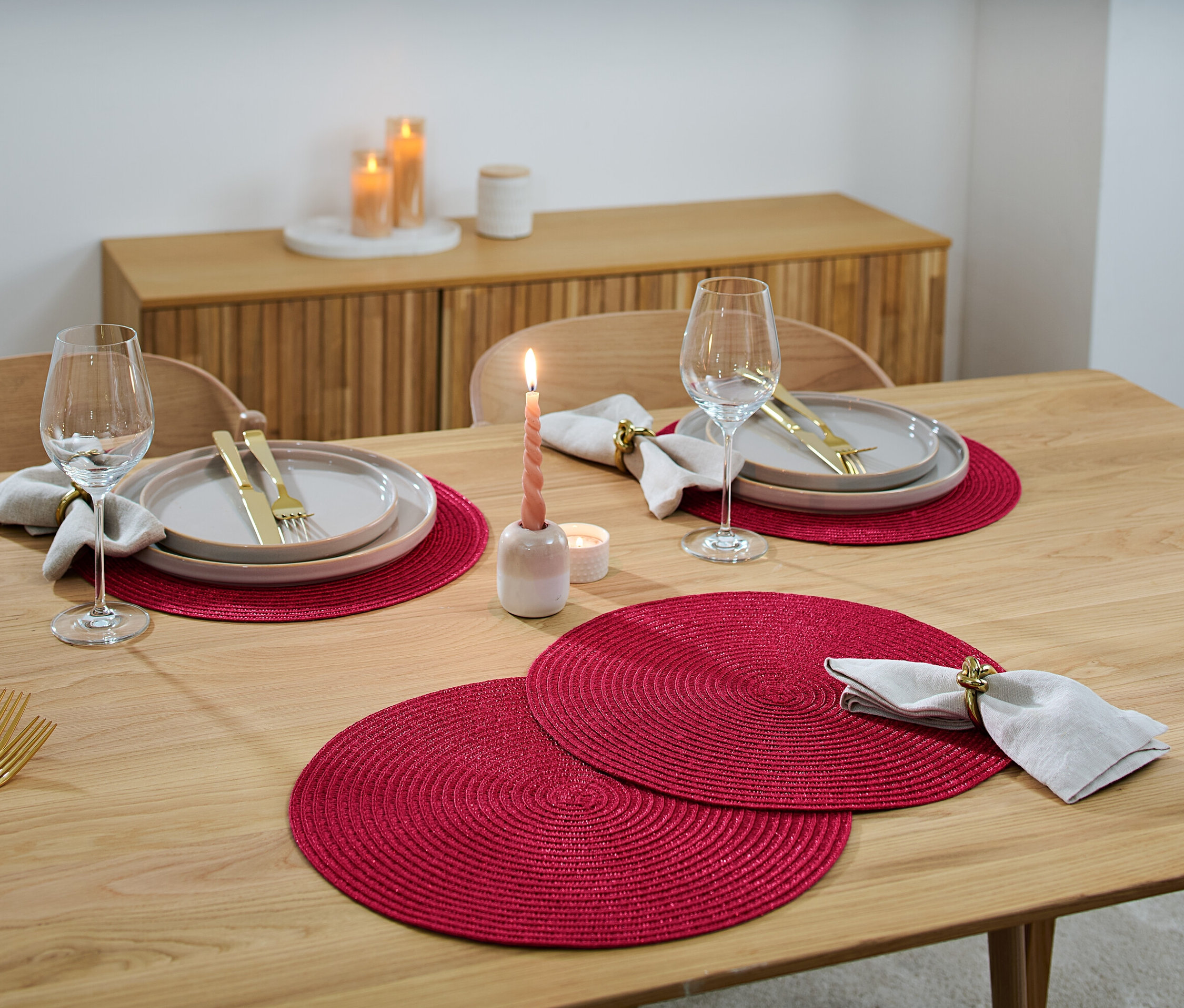 Sur une table en bois sont disposés des couverts avec 4 sets de table, des assiettes, des verres à vin, des couverts et des bougies.