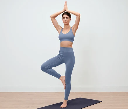 Femme en Legging Seamless bleu debout sur un tapis de yoga dans la pose de l'arbre.