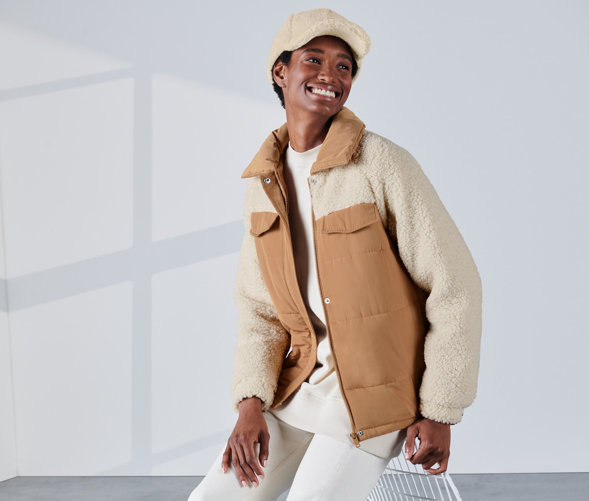 Une femme souriante est assise et porte une veste matelassée de couleur cognac et blanche avec des accents en teddy et un béret.