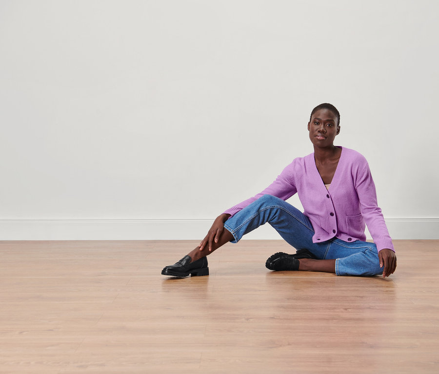 Une femme est assise par terre et porte une jaquette en maille, un jean et des chaussures.