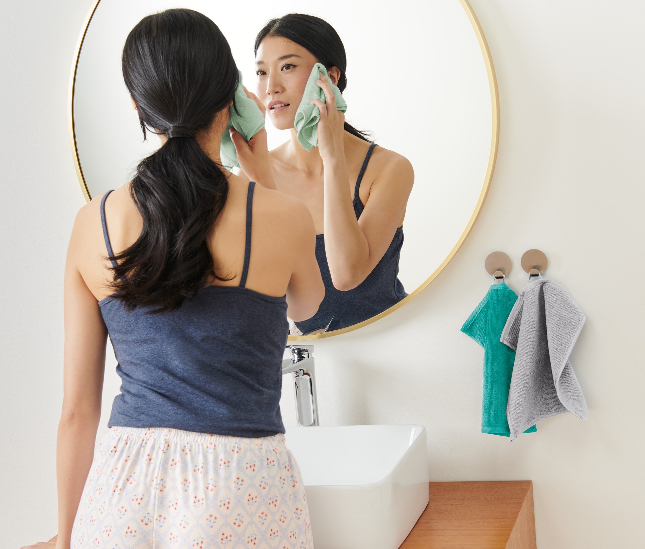 Une femme se nettoie le visage avec un lot de 3 lingettes cosmétiques en microfibre devant un miroir.