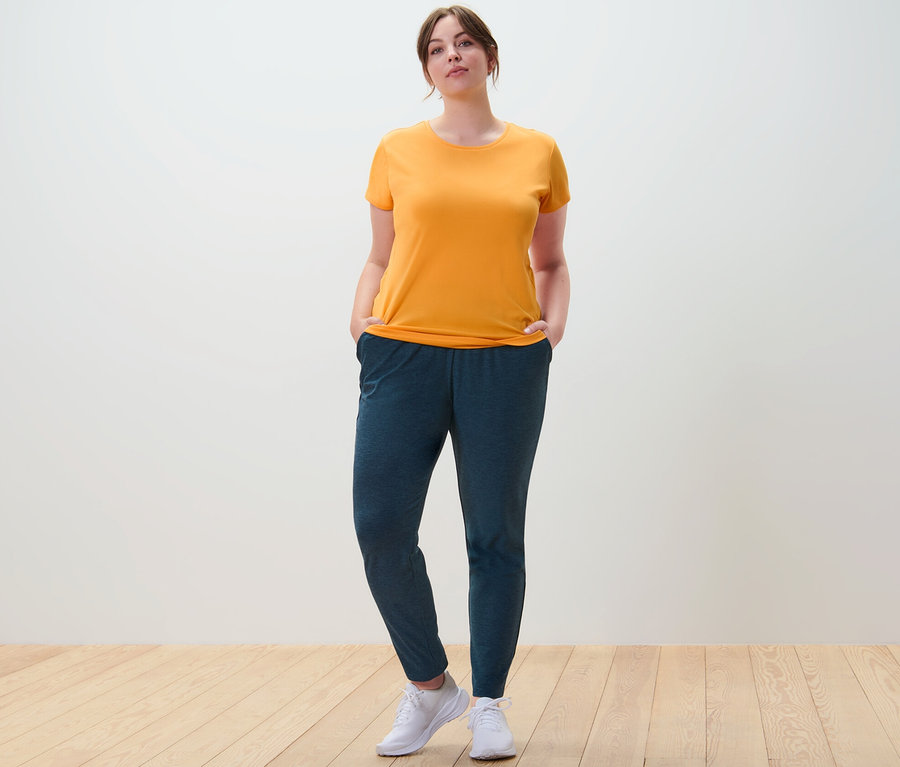 Une femme se tient debout, les mains dans les poches de son pantalon de sport, portant un t-shirt technique orange.