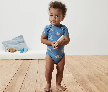 Un bébé se tient debout et tient un bloc de bois, il porte un body bleu à manches courtes.