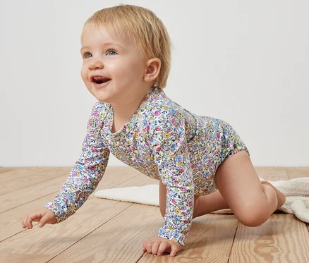Un bébé rampe sur un plancher en bois et porte un body rose à fleurs.