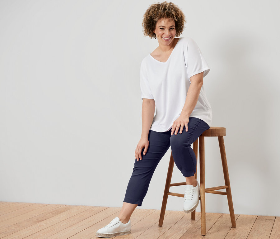 Une femme est assise sur un tabouret et porte une blouse structurée blanche et un pantalon stretch 7/8 bleu foncé.