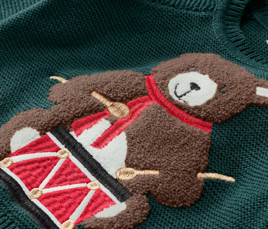 Gros plan sur un pull vert avec un appliqué d'ours en peluche jouant du tambour.