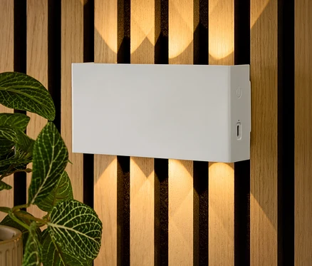Une applique murale rectangulaire blanche est fixée à un mur en bois. En dessous, des clés sont posées sur une table et une fleur est dans un pot.