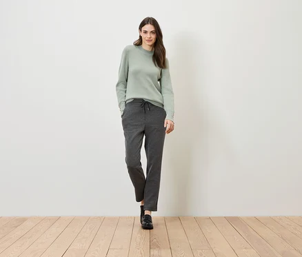 Une femme se tient sur un plancher en bois devant un mur blanc. Elle porte un pull vert et un pantalon gris.