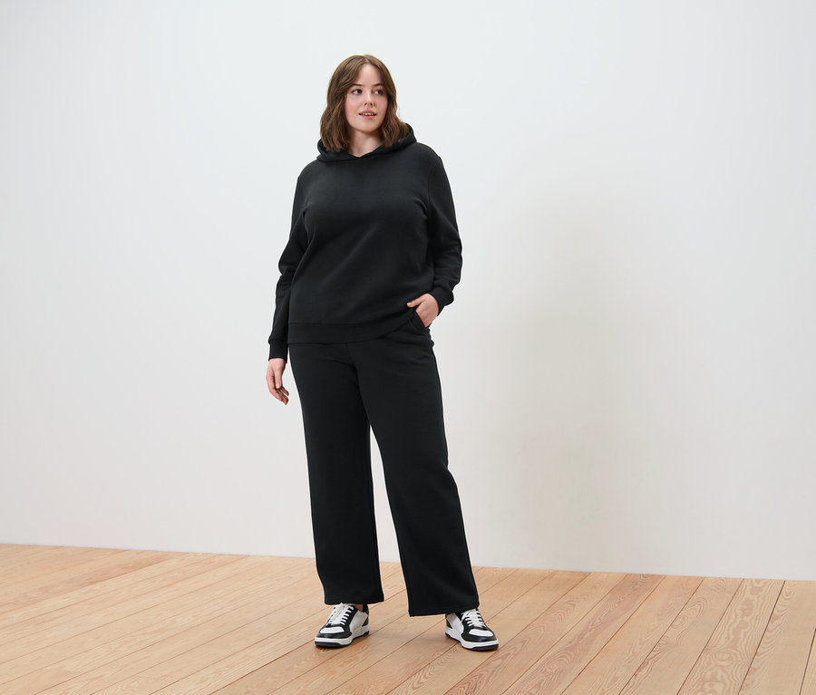Femme portant un ensemble sweat à capuche noir avec un pantalon large, debout sur un plancher en bois.