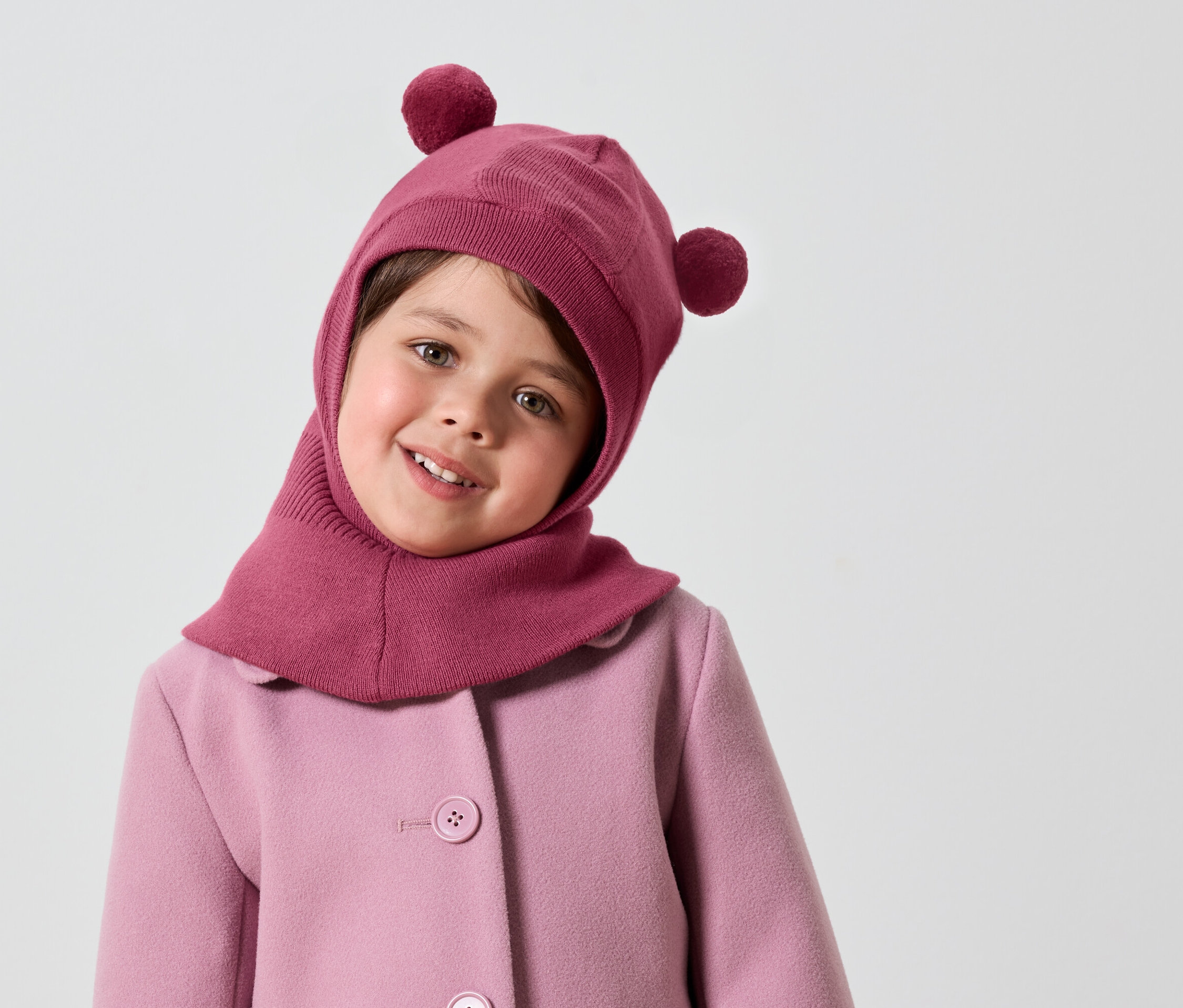 Fillette souriante portant une cagoule rose à pompons et un manteau rose.