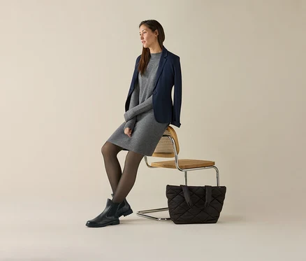 Une femme est assise sur une chaise et porte une robe grise et un blazer en jersey bleu marine. Un sac à main est posé à côté de la chaise.