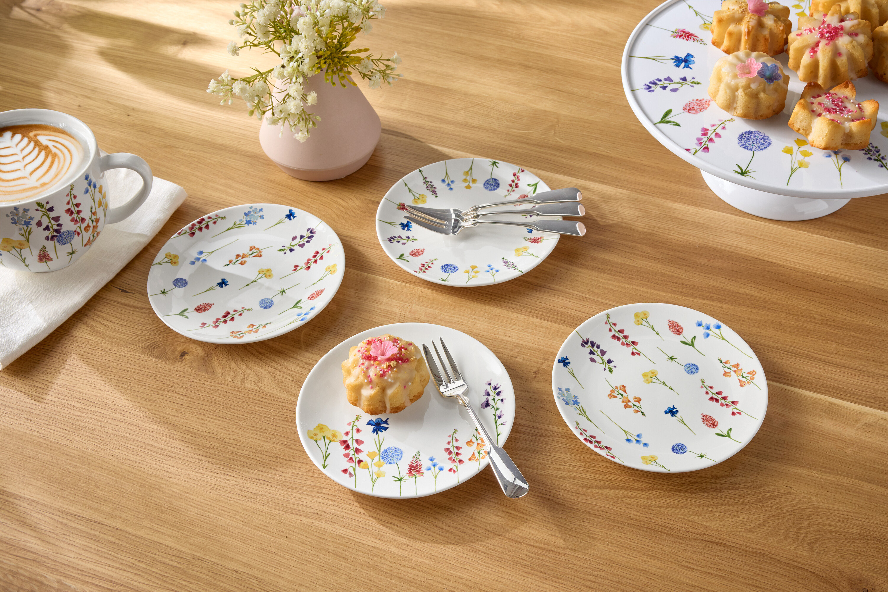 Sur une table en bois, il y a une tasse de café, un vase de fleurs, des assiettes et des petits gâteaux.