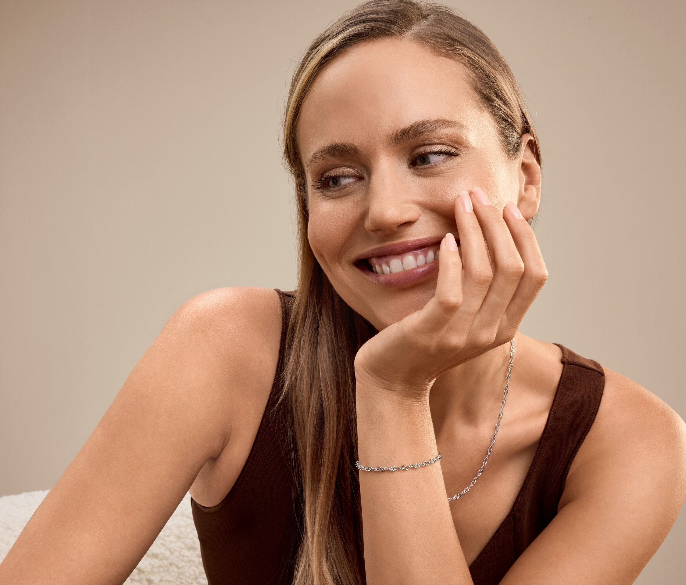 Une femme blonde souriante, le visage appuyé sur sa main, porte un bracelet et un collier.