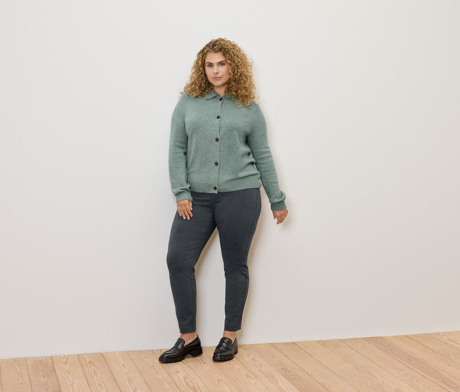 Une femme aux cheveux bouclés portant un cardigan vert, un pantalon gris et des mocassins noirs, debout.