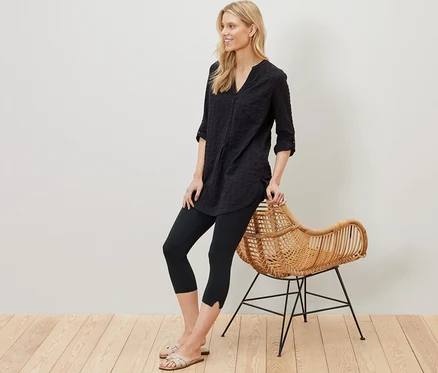 Une femme blonde appuiyée sur un fauteuil en rotin porte un legging 3/4 noir avec détail.
