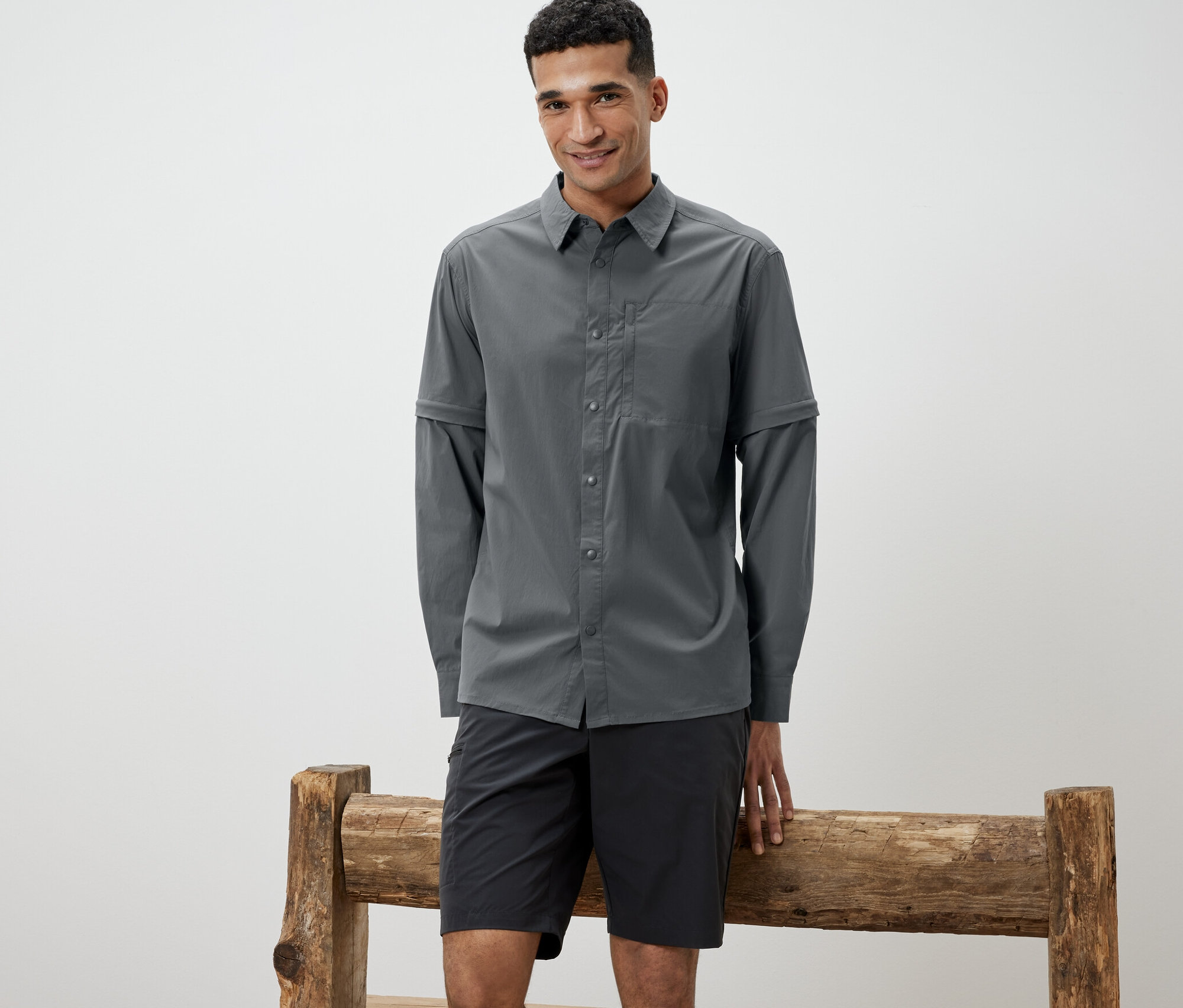 Un homme appuyé contre un banc en bois porte une chemise d’extérieur grise et un short noir.
